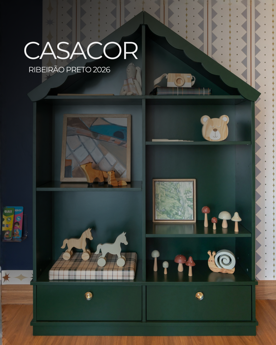 Estante Fábula em verde escuro, em formato de casinha, com telhado recortado, prateleiras abertas e duas gavetas na base.